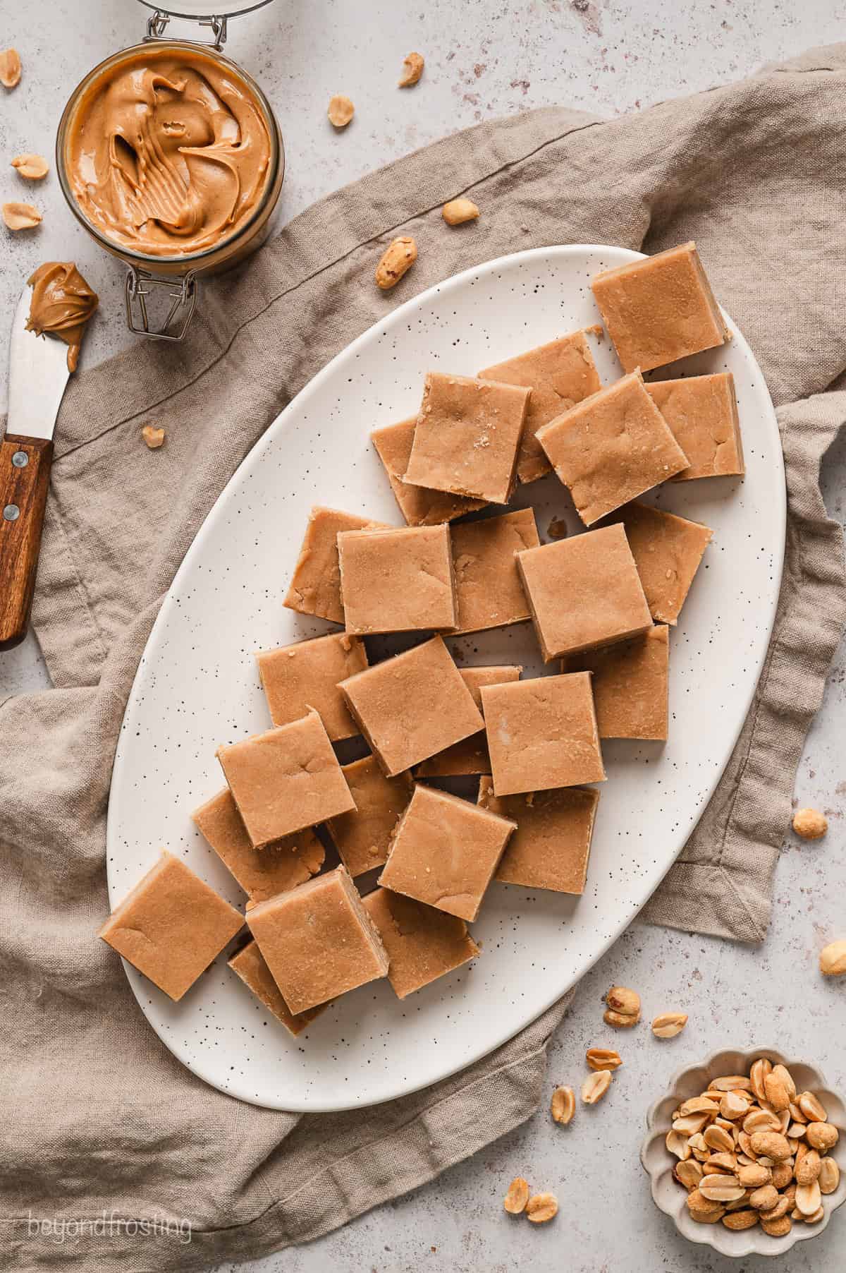 Overhead view of peanut butter fudge served on a large oval platter, next to a jar of peanut butter and a bowl of peanuts.