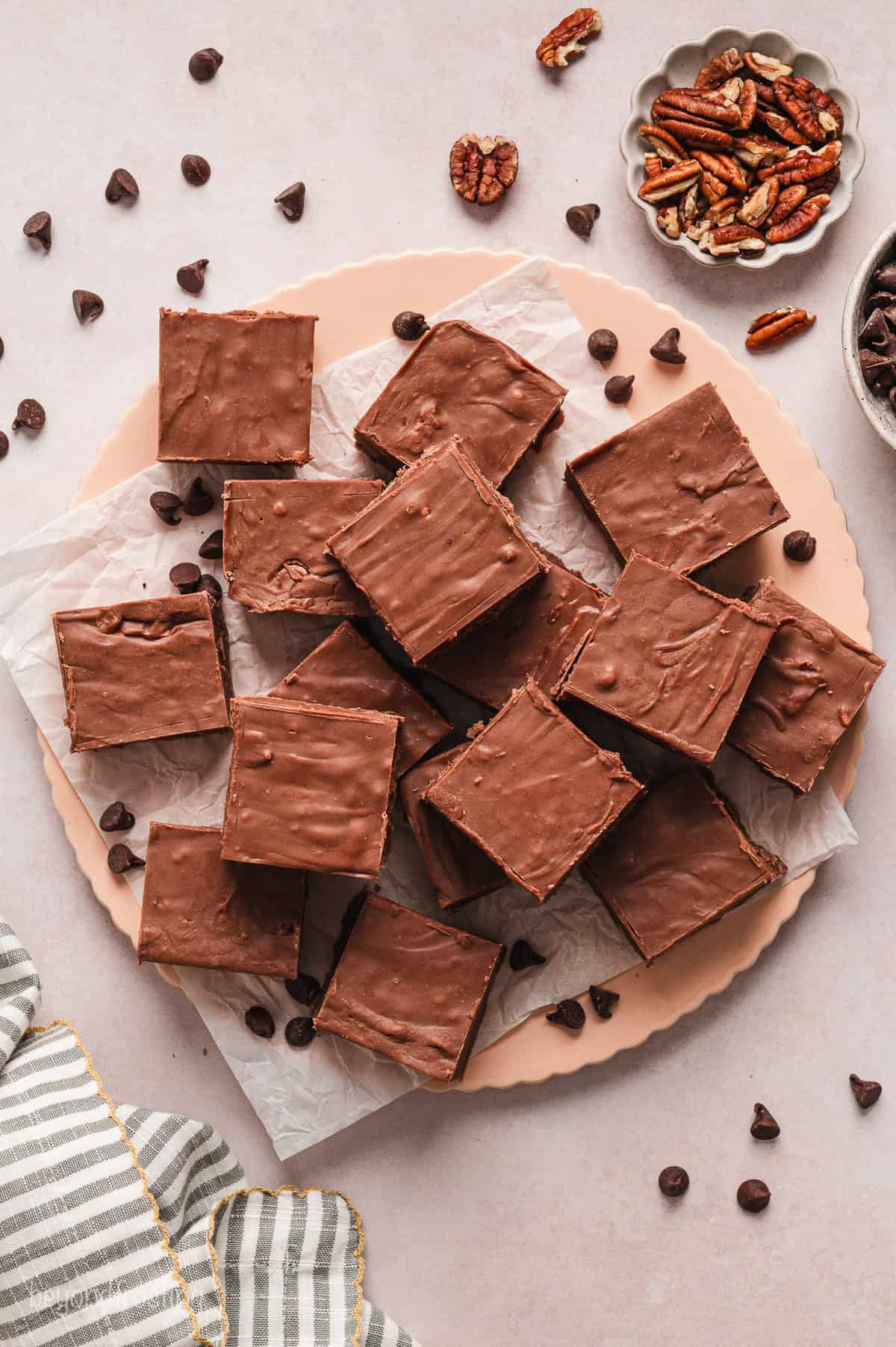 Overhead view of fantasy fudge squares stacked on a parchment paper-lined wooden platter, surrounded by scattered chocolate chips and pecans.