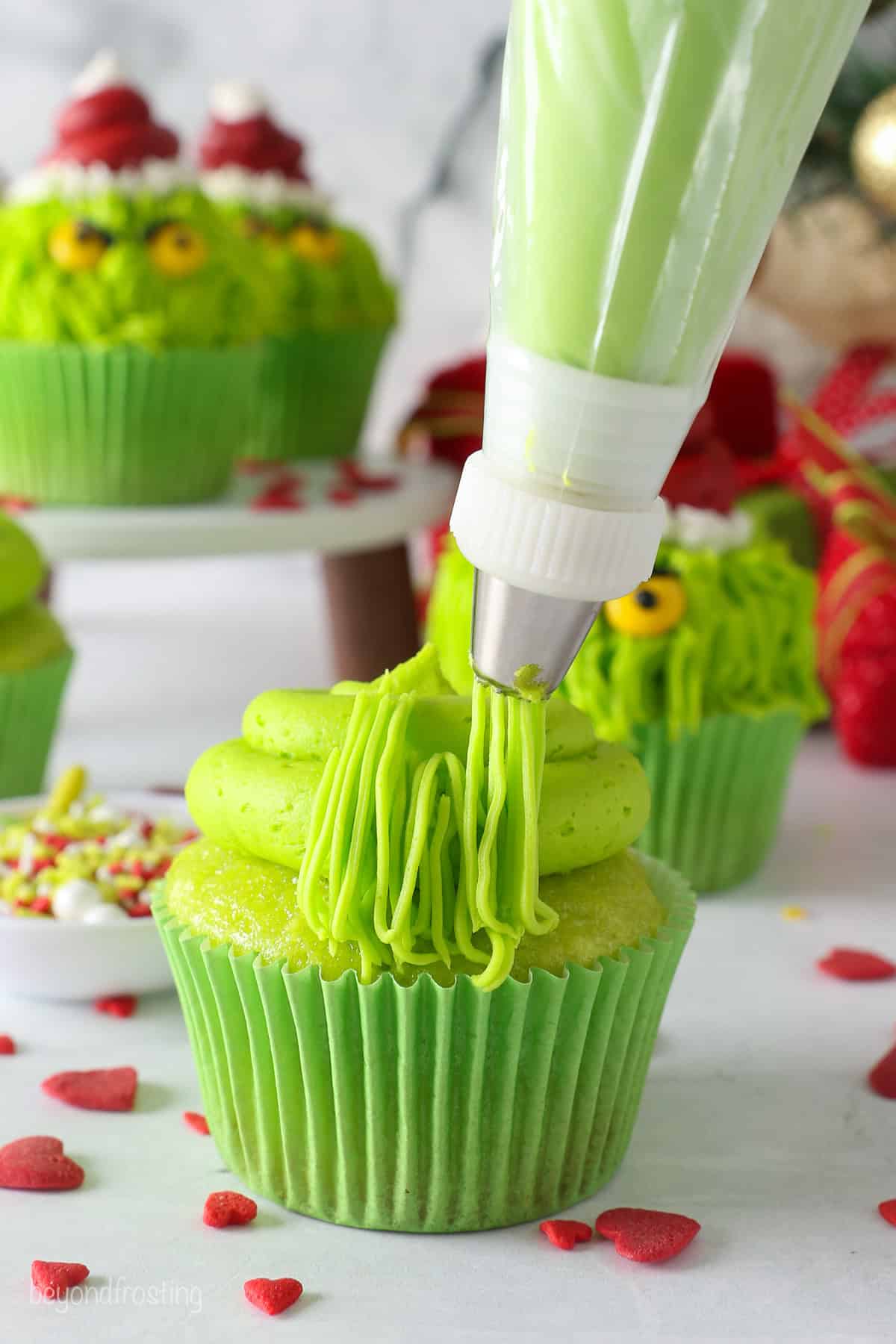 A piping bag fitted with a grass tip piping green frosting hair over a swirl of green frosting on a cupcake.