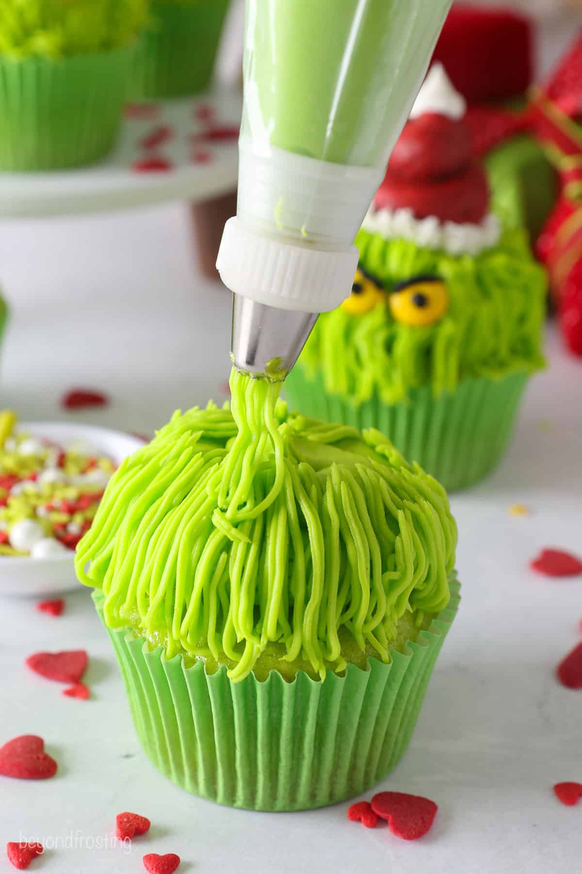 A piping bag fitted with a grass tip piping green frosting hair over a green cupcake.