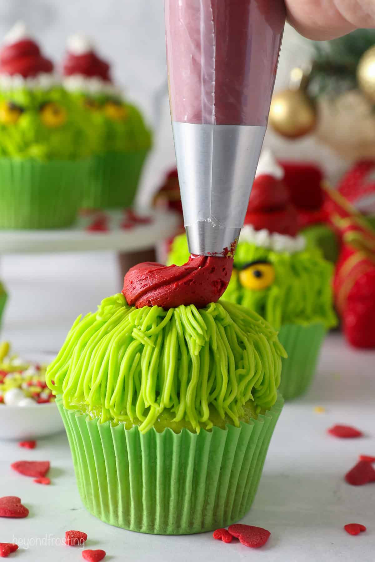 A piping bag piping red frosting in the shape of a Santa hat over a frosted green Grinch cupcake.