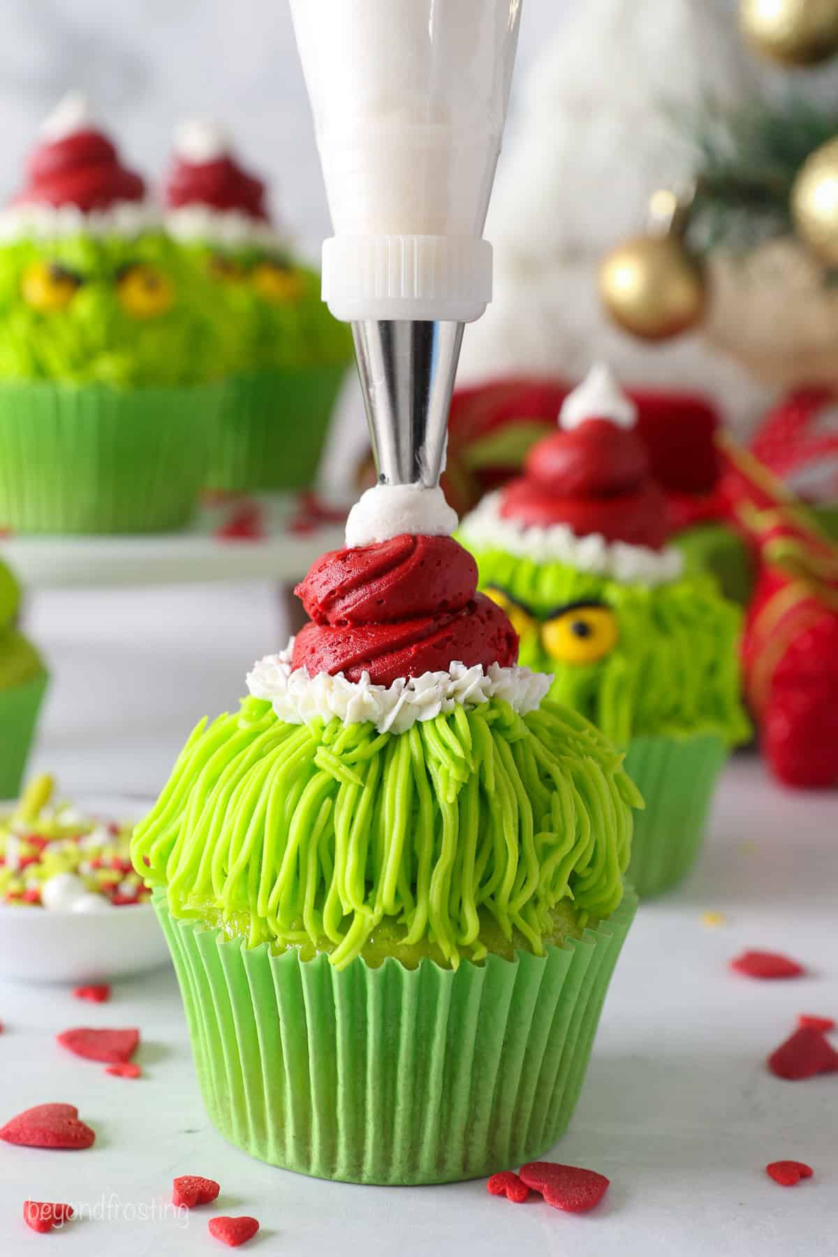 A dollop of white frosting being piped on the tip of the red frosting Santa hat on a green Grinch cupcake.