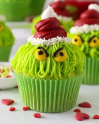 A Grinch cupcake decorated with a red frosting Santa hat, yellow M&M eyes, and black frosting details, with more Grinch cupcakes in the background.