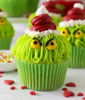 A Grinch cupcake decorated with a red frosting Santa hat, yellow M&M eyes, and black frosting details, with more Grinch cupcakes in the background.