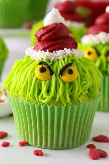 A Grinch cupcake decorated with a red frosting Santa hat, yellow M&M eyes, and black frosting details, with more Grinch cupcakes in the background.