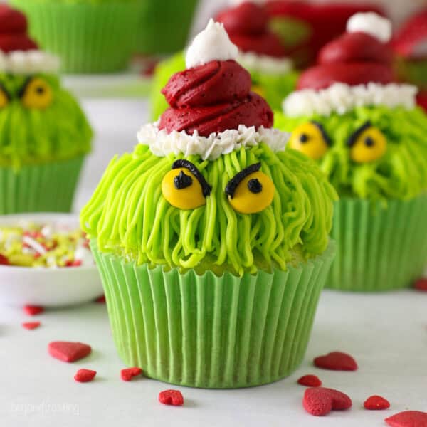 A Grinch cupcake decorated with a red frosting Santa hat, yellow M&M eyes, and black frosting details, with more Grinch cupcakes in the background.