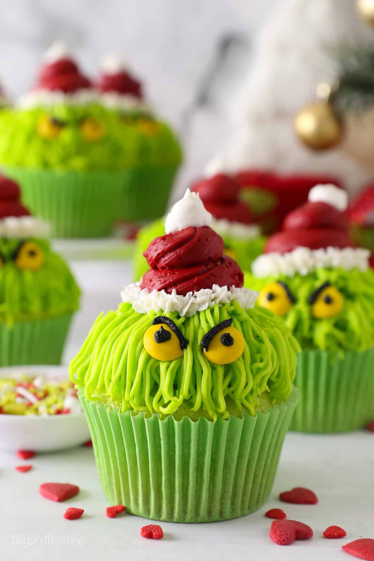 Grinch cupcakes decorated with red frosting Santa hats, yellow M&M eyes, and black frosting details, with more cupcakes in the background.