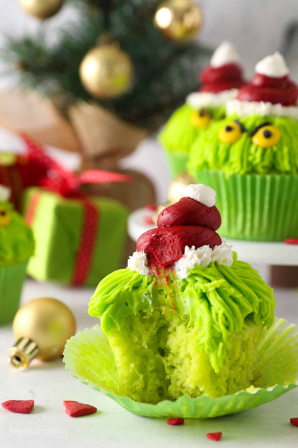 A Grinch cupcake unwrapped with a bite missing, with more cupcakes on a raised cake stand in the background.