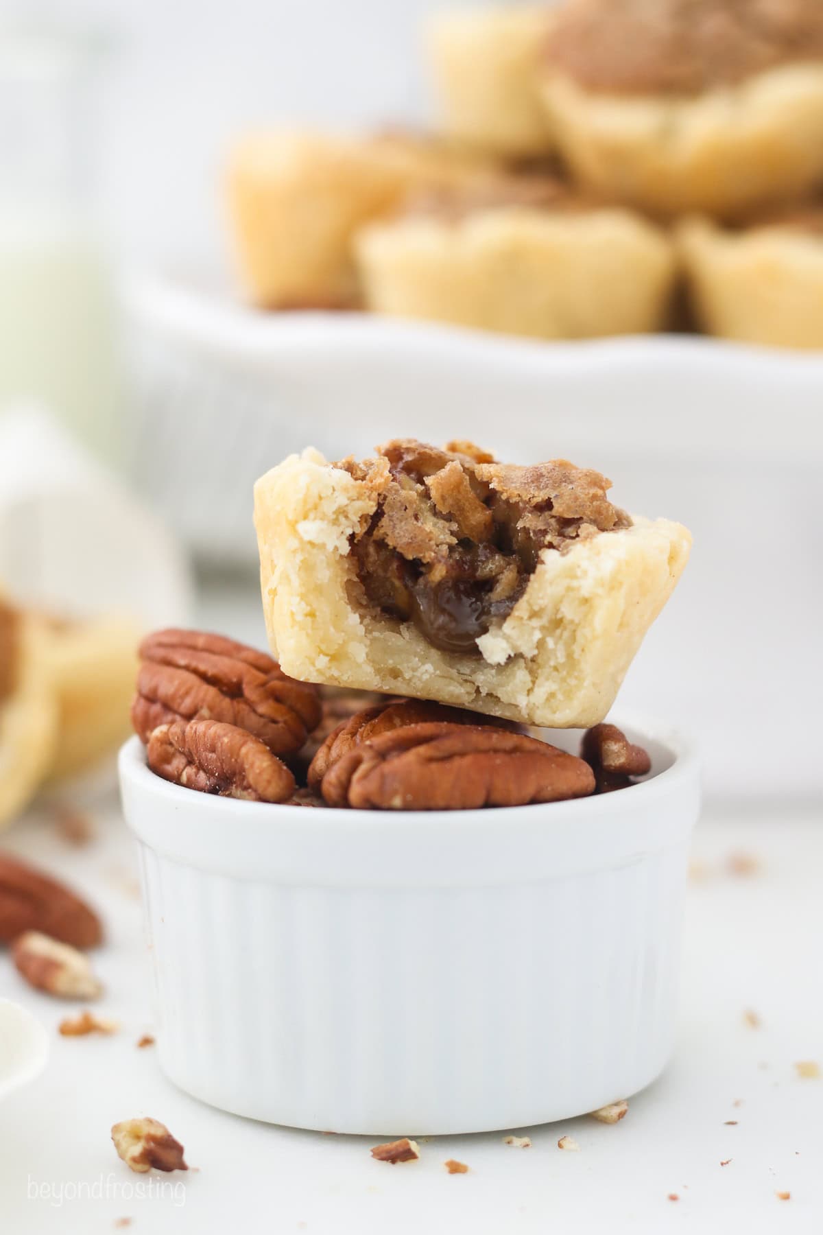 A mini pecan tart with a bite missing stacked on top of a small ramekin filled with pecans.