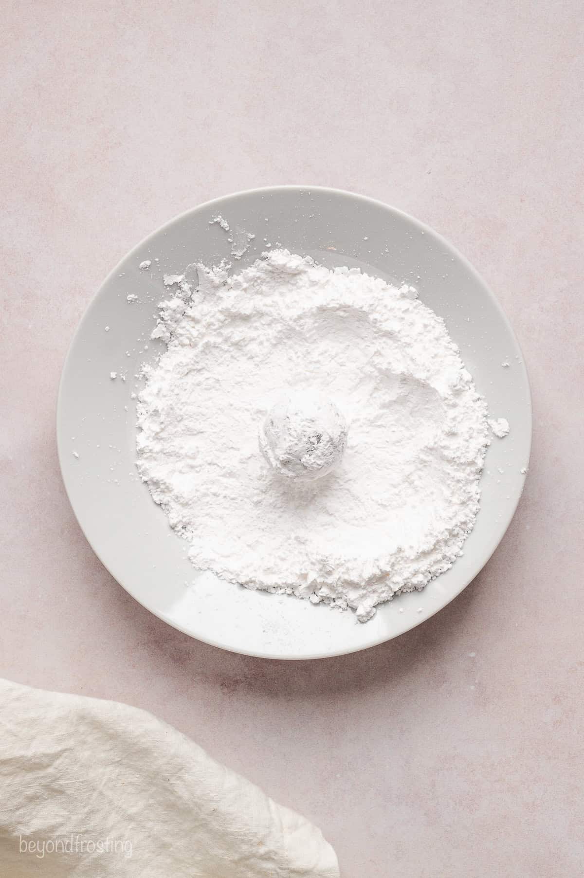 A sugar-coated chocolate cookie dough ball resting in a bowl of powdered sugar.