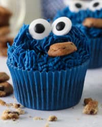 Close up of a Cookie Monster cupcake surrounded by scattered mini Chips Ahoy cookies, with more cupcakes in the background.