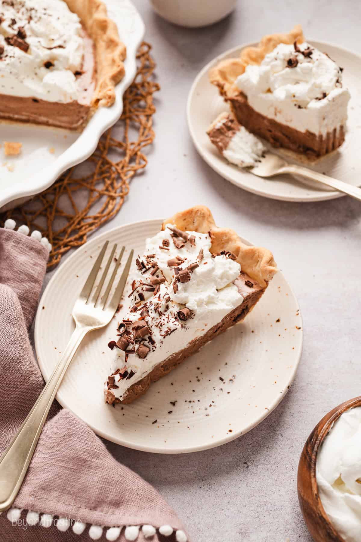 A slice of French silk pie on a plate next to a fork, with the rest of the pie and a second partially eaten slice in the background.