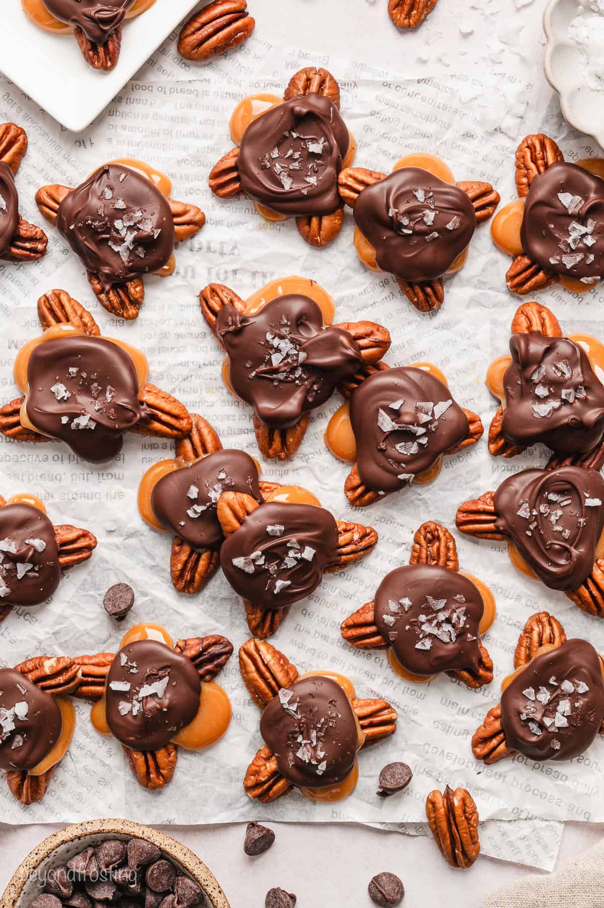 Overhead view of scattered turtle candies topped with flaky sea salt on a sheet of parchment paper.