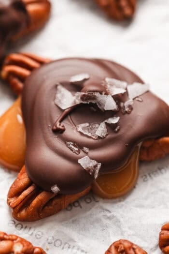 Close up of turtle candies topped with flaky sea salt on a sheet of parchment paper.