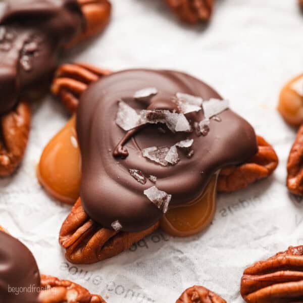 Close up of turtle candies topped with flaky sea salt on a sheet of parchment paper.