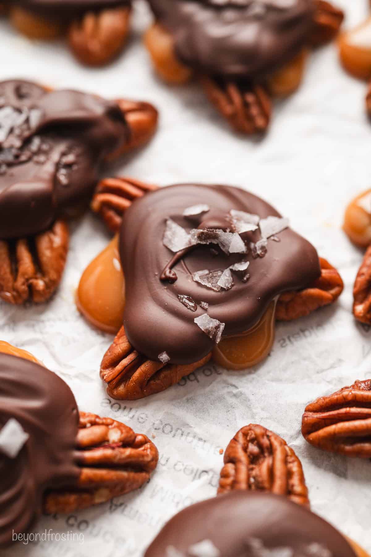 Close up of turtle candies topped with flaky sea salt on a sheet of parchment paper.