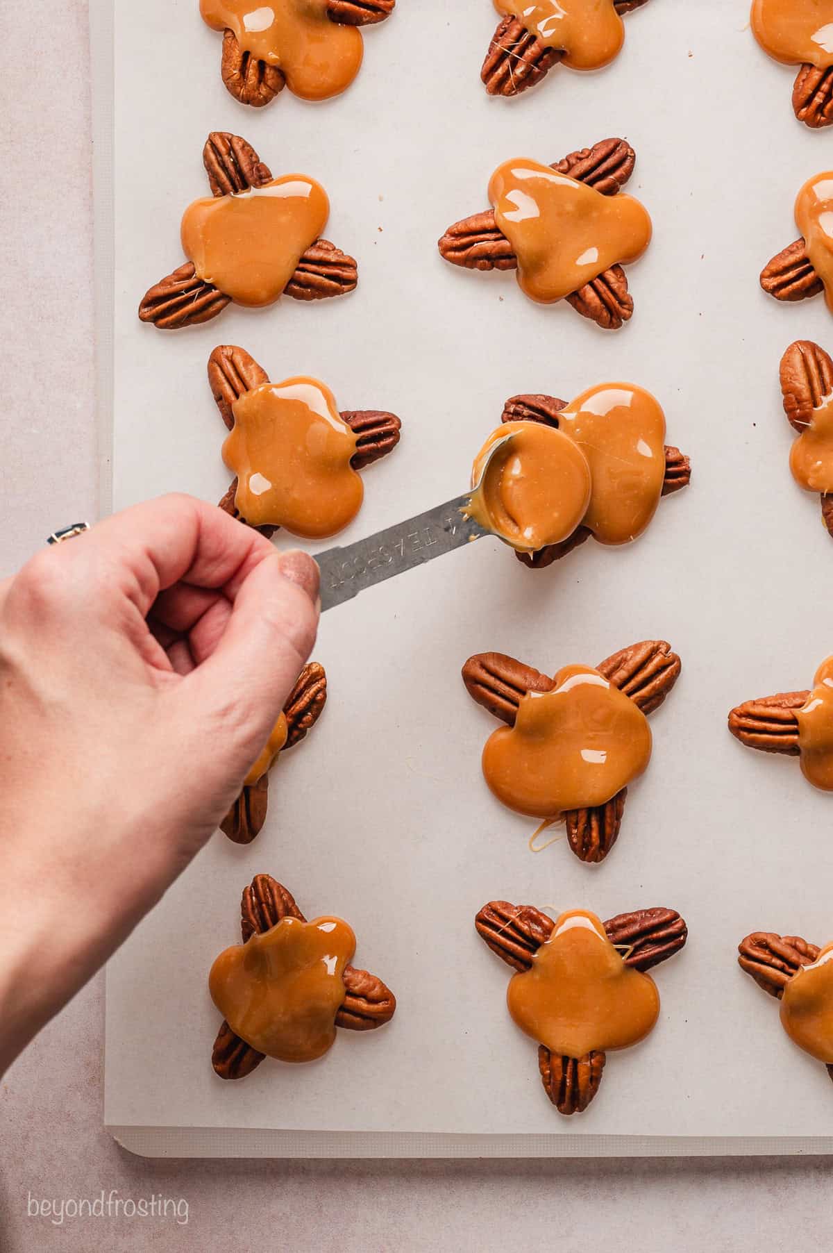 A hand spooning melted caramel over pecan clusters on a sheet of parchment paper.