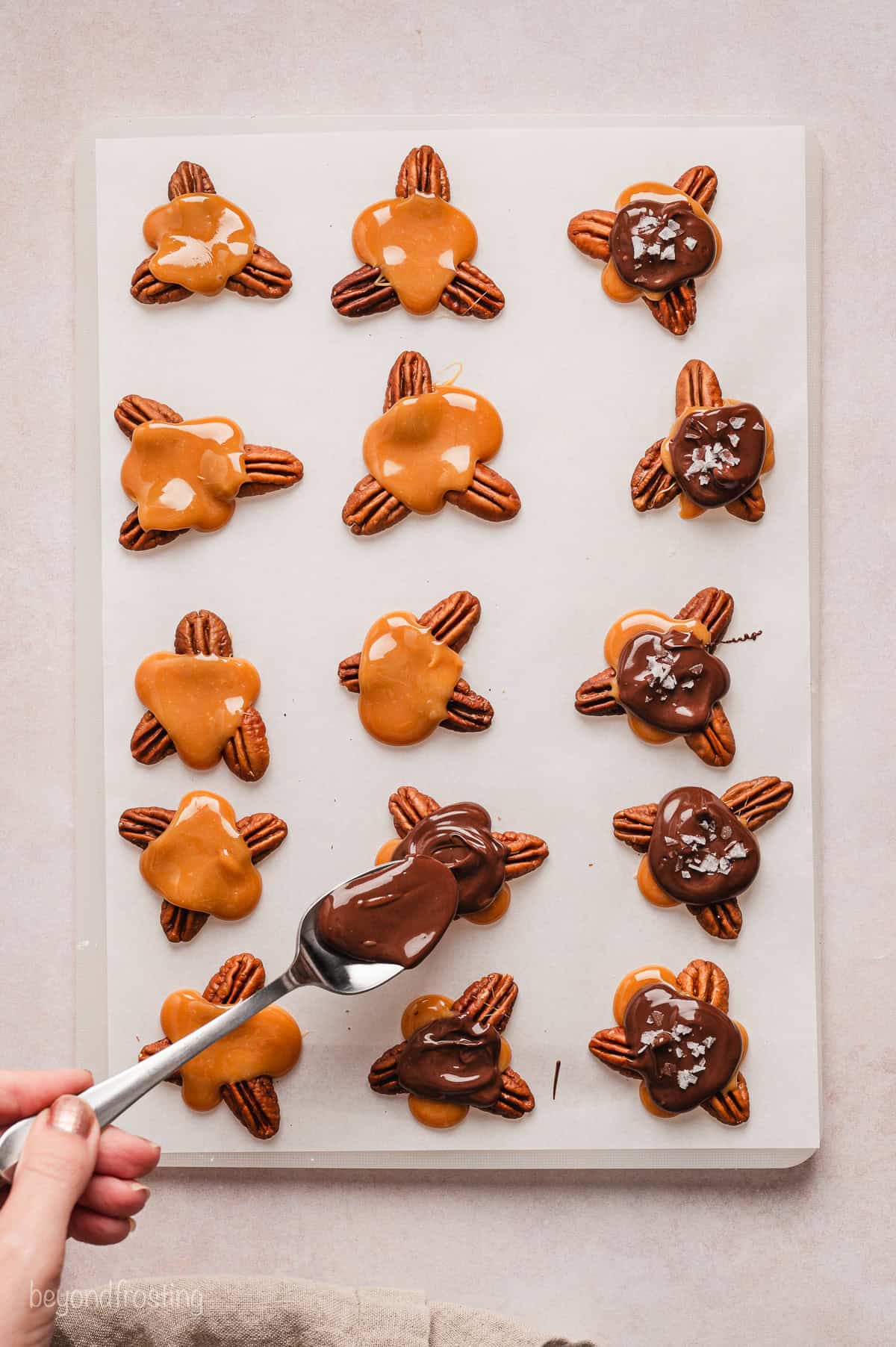 A hand spooning melted chocolate over partially assembled turtle candies on a sheet of parchment paper.