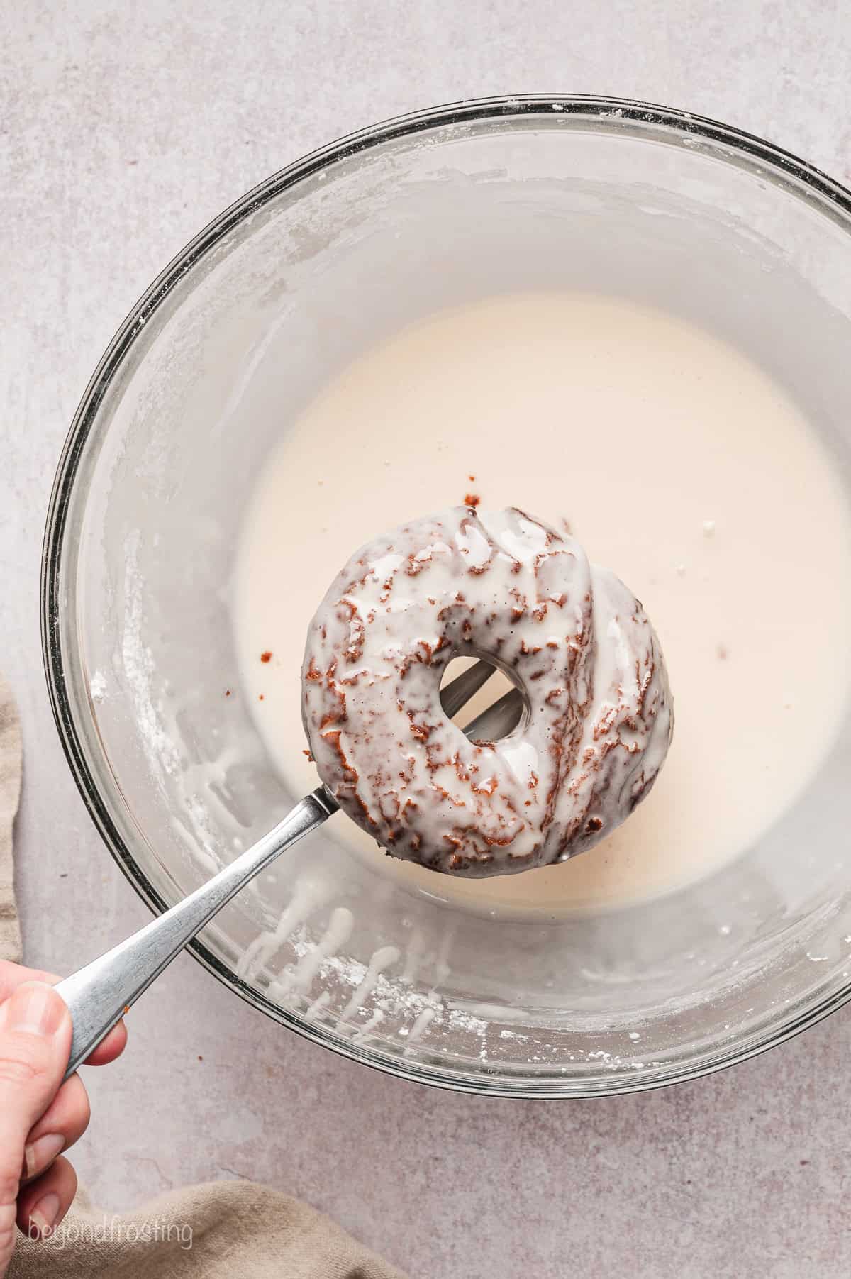 A glazed chocolate donut held over a bowl of glaze with a fork.