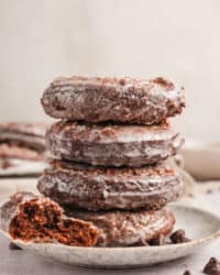 Half of a chocolate donut resting next to a stack of four glazed donuts on a plate.