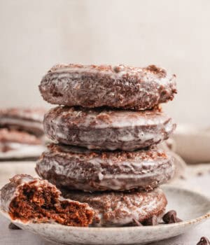 Half of a chocolate donut resting next to a stack of four glazed donuts on a plate.