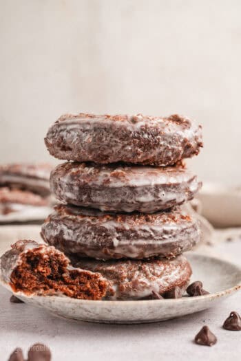 Half of a chocolate donut resting next to a stack of four glazed donuts on a plate.