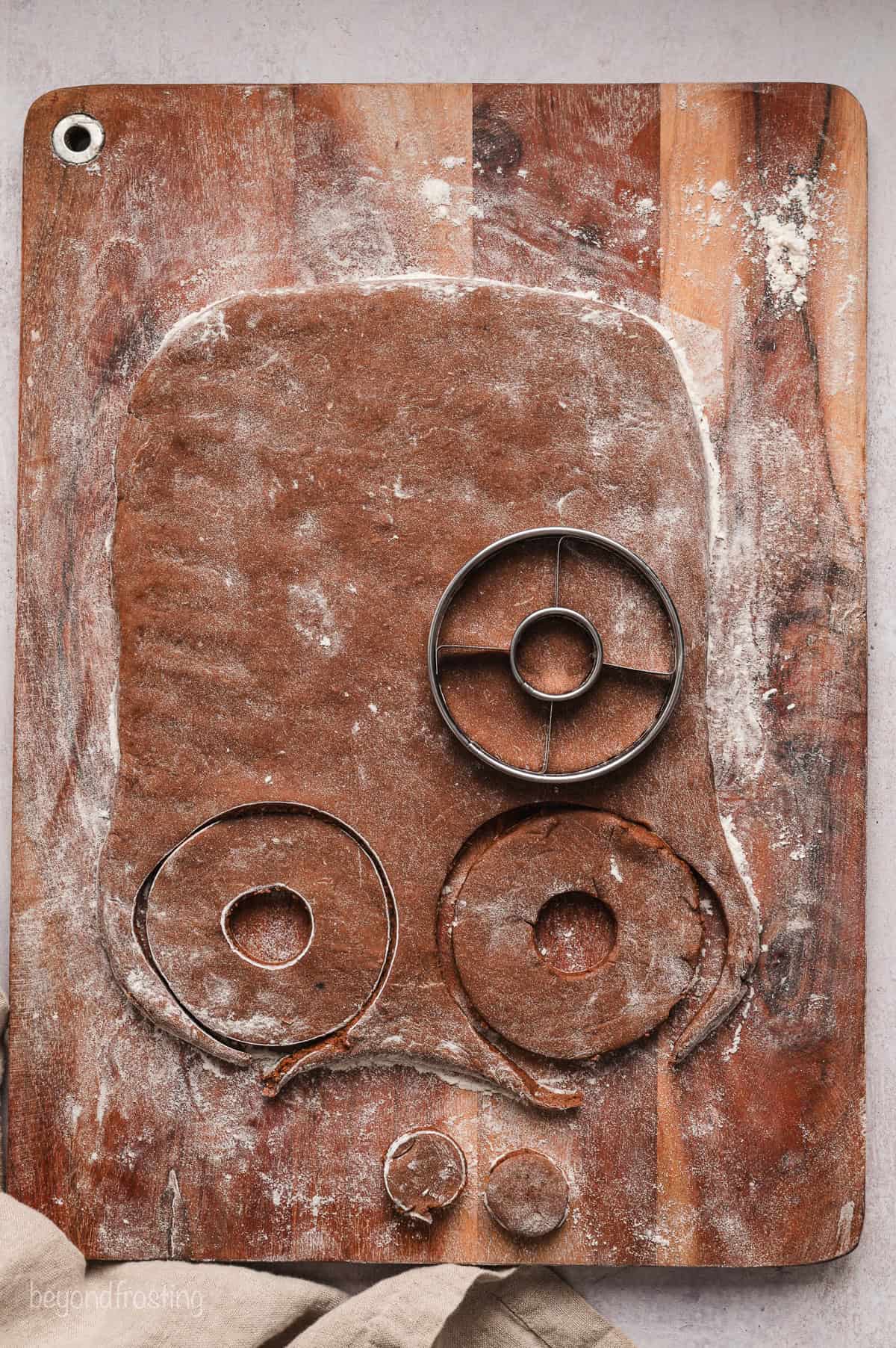 A donut cutter being used to cut out donuts from rolled out chocolate donut dough on a wooden cutting board.