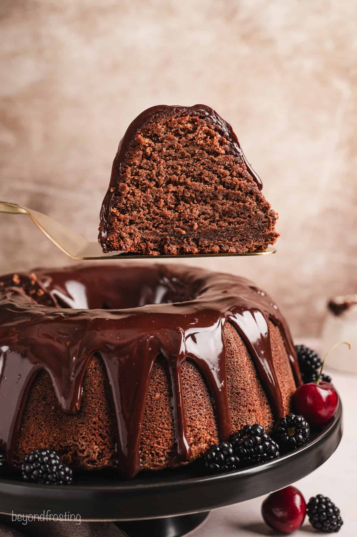 A slice of chocolate pound cake being lifted with a cake server above the rest of the glazed pound cake on a cake stand.