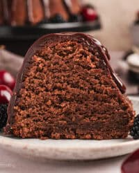 A slice of glazed chocolate pound cake on a plate next to cherries and blackberries, with the rest of the pound cake on a cake stand in the background.