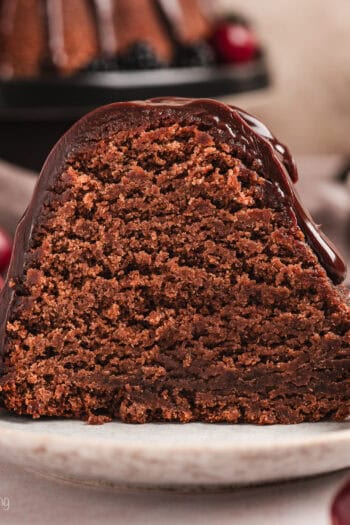 A slice of glazed chocolate pound cake on a plate next to cherries and blackberries, with the rest of the pound cake on a cake stand in the background.