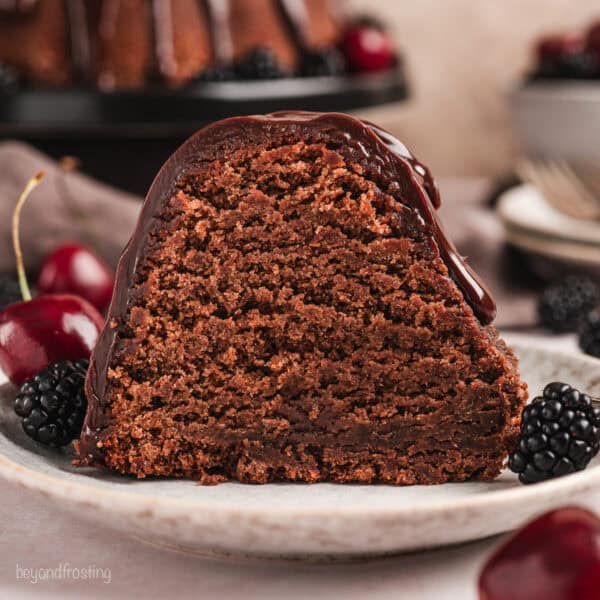 A slice of glazed chocolate pound cake on a plate next to cherries and blackberries, with the rest of the pound cake on a cake stand in the background.