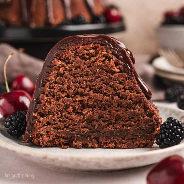 A slice of glazed chocolate pound cake on a plate next to cherries and blackberries, with the rest of the pound cake on a cake stand in the background.