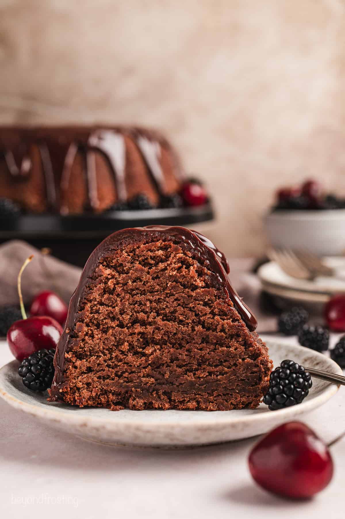 A slice of glazed chocolate pound cake on a plate next to cherries and blackberries, with the rest of the pound cake on a cake stand in the background.