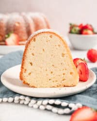 A slice of cream cheese pound cake on a plate next to fresh strawberries, with a bowl of strawberries and the rest of the cake in the background.