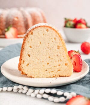 A slice of cream cheese pound cake on a plate next to fresh strawberries, with a bowl of strawberries and the rest of the cake in the background.