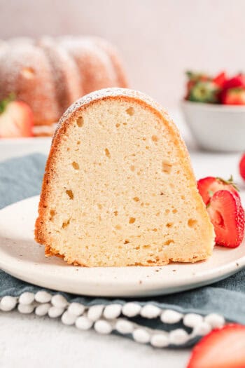 A slice of cream cheese pound cake on a plate next to fresh strawberries, with a bowl of strawberries and the rest of the cake in the background.