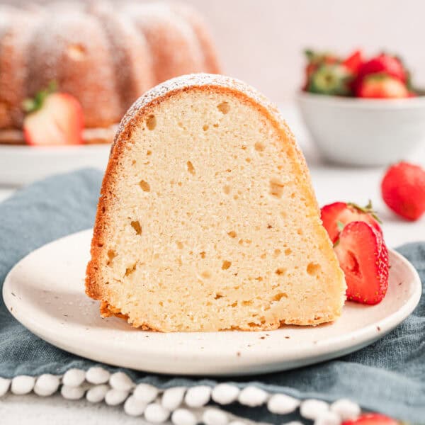 A slice of cream cheese pound cake on a plate next to fresh strawberries, with a bowl of strawberries and the rest of the cake in the background.