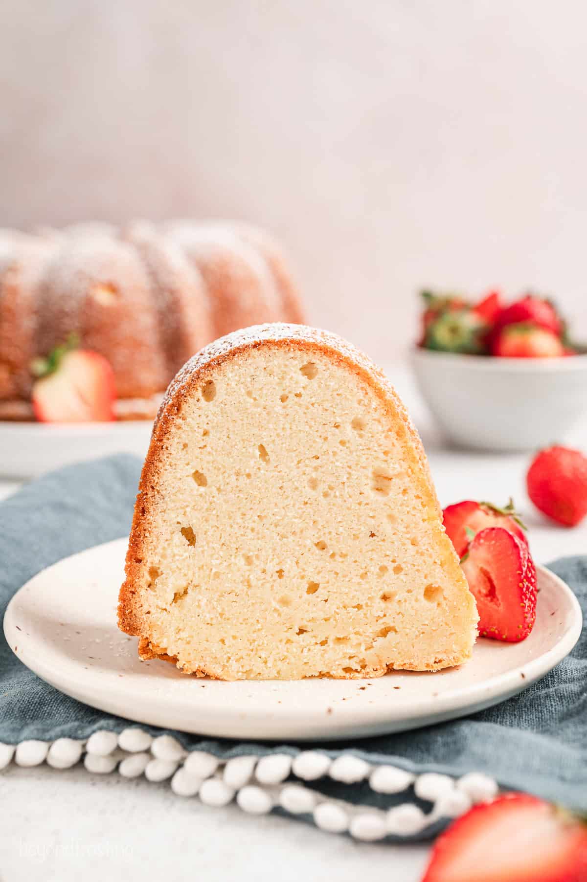 A slice of cream cheese pound cake on a plate next to fresh strawberries, with a bowl of strawberries and the rest of the cake in the background.