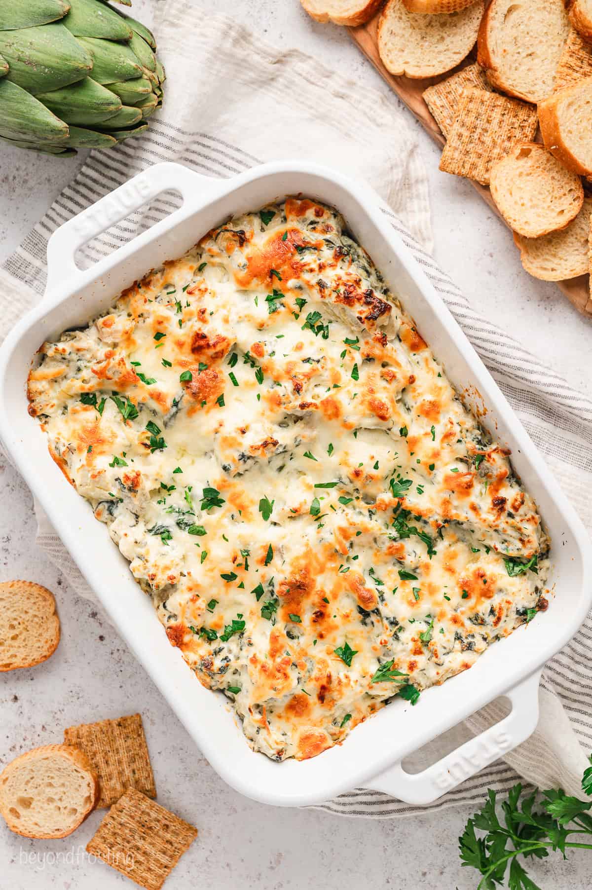 Overhead view of baked spinach artichoke dip in a ceramic dish next to a platter of crostini.