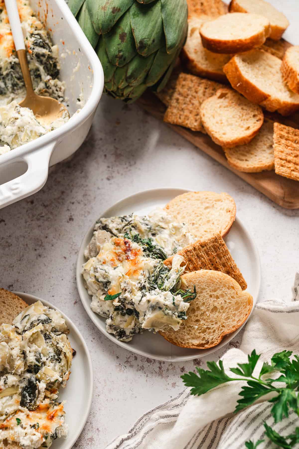 Overhead view of crostini next to spinach artichoke dip on a plate, next to more crostini on a platter and artichoke dip in a baking dish.