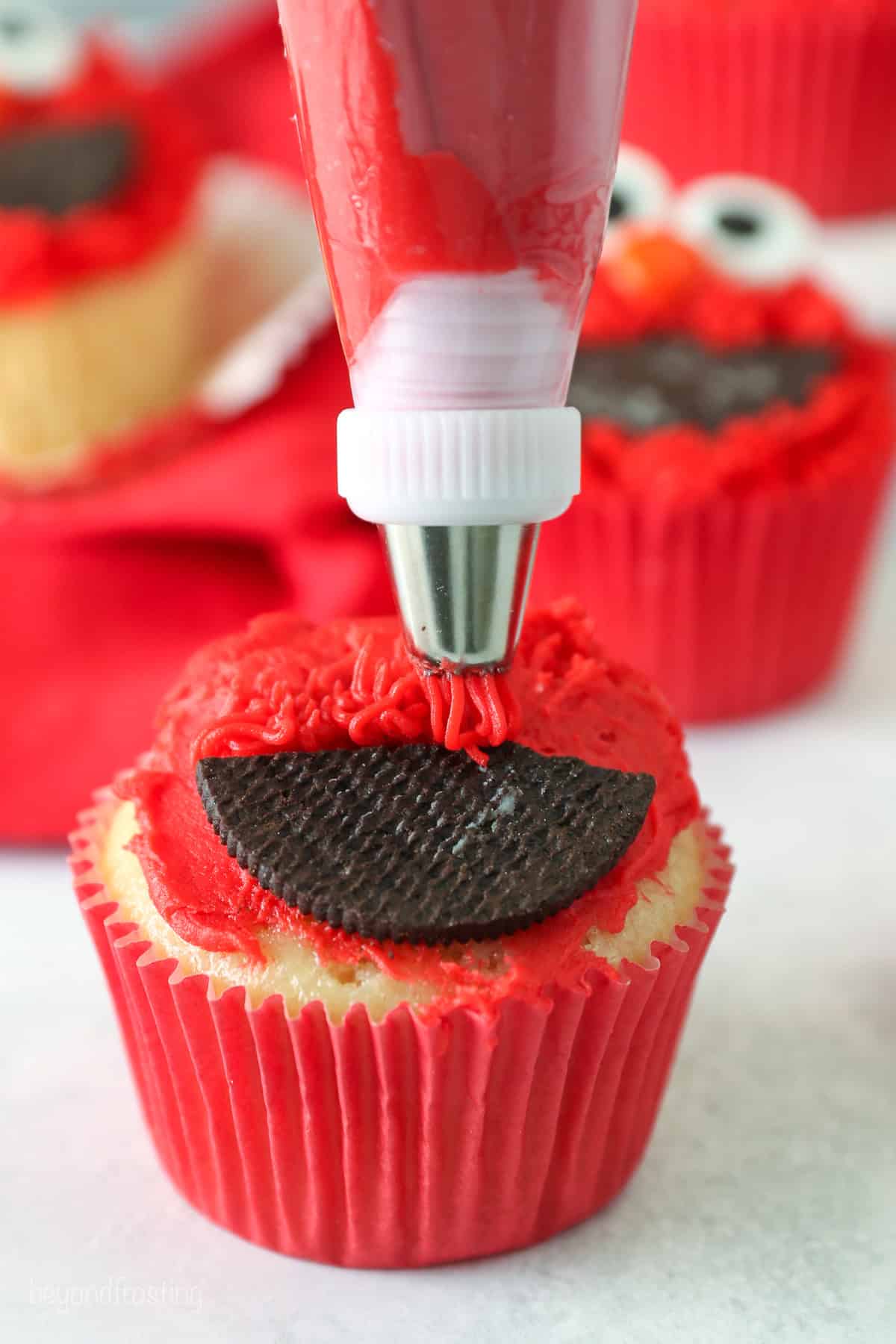 A piping tip pipes red frosting fur over a partially decorated Elmo cupcake.