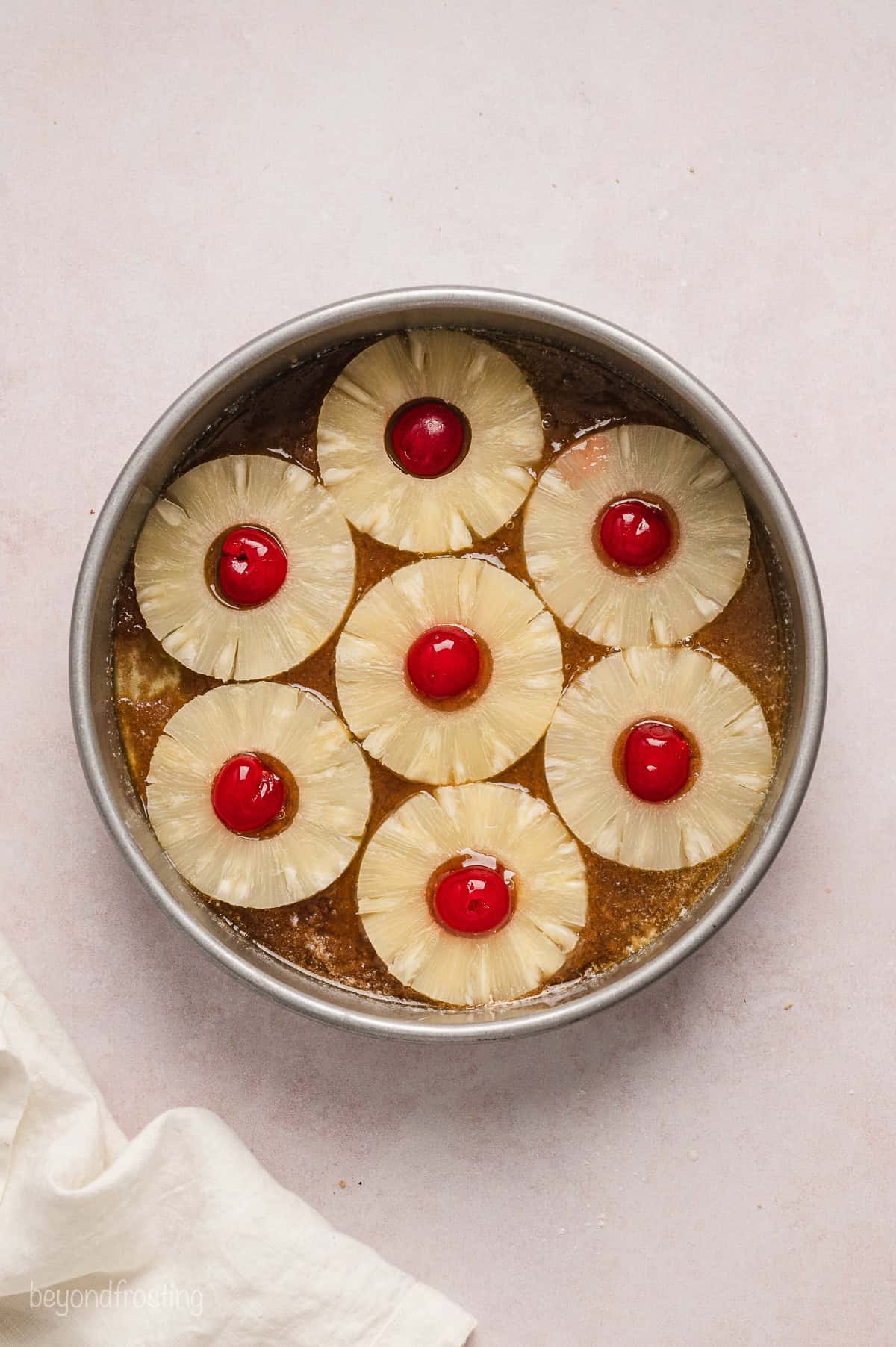 Pineapple rings and maraschino cherries arranged over top of the brown sugar base inside a round cake pan.