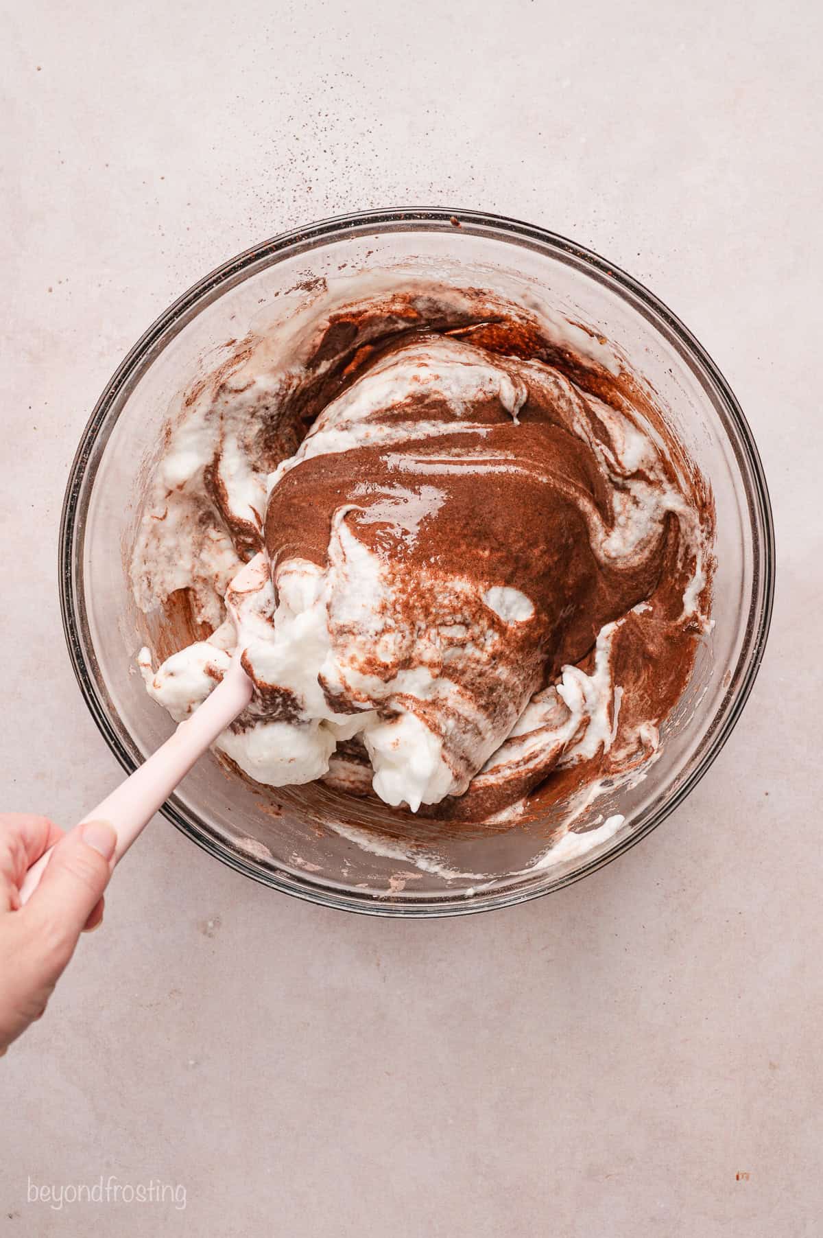 A hand using a spatula to fold egg whites into chocolate cake batter in a glass bowl.