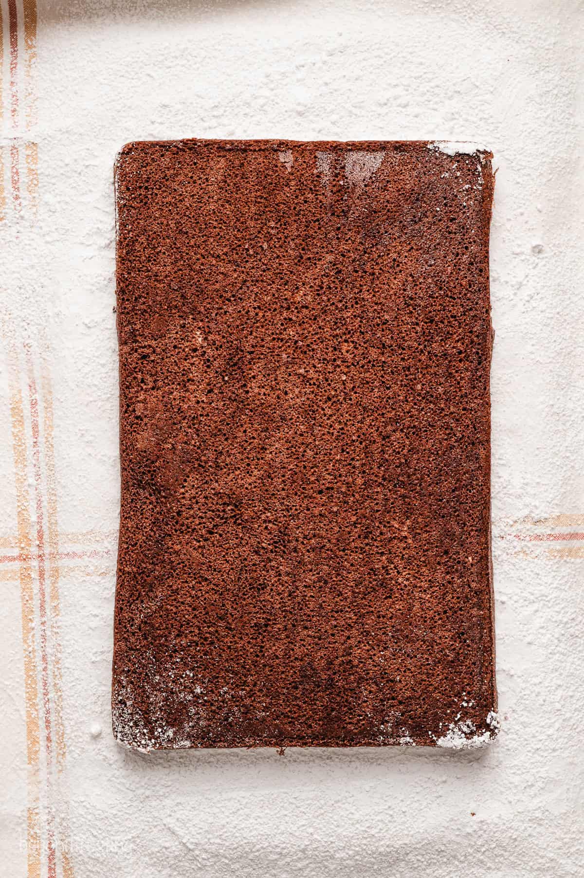 A sheet of chocolate cake on top of a clean dish towel covered with powdered sugar.