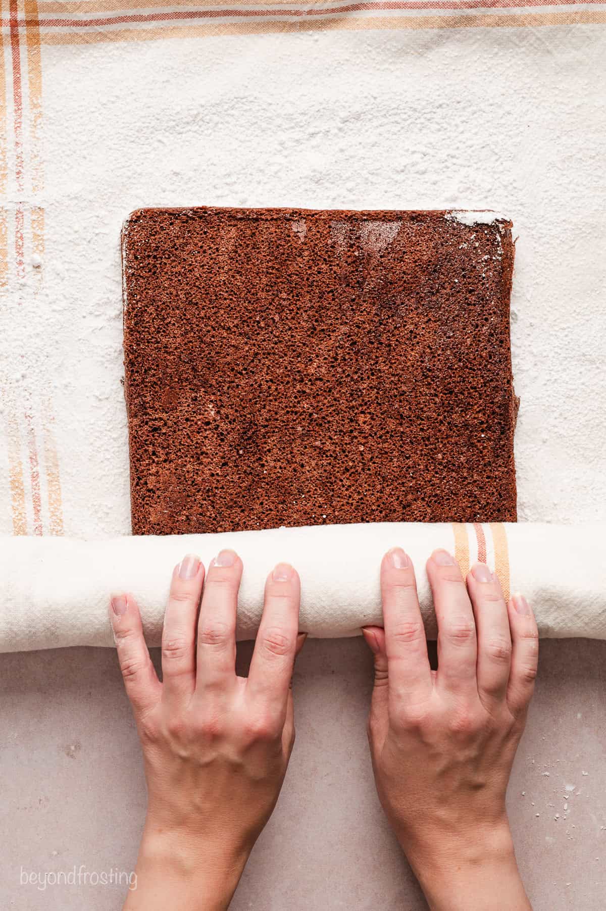 Two hands rolling a sheet of chocolate cake inside a dish towel.