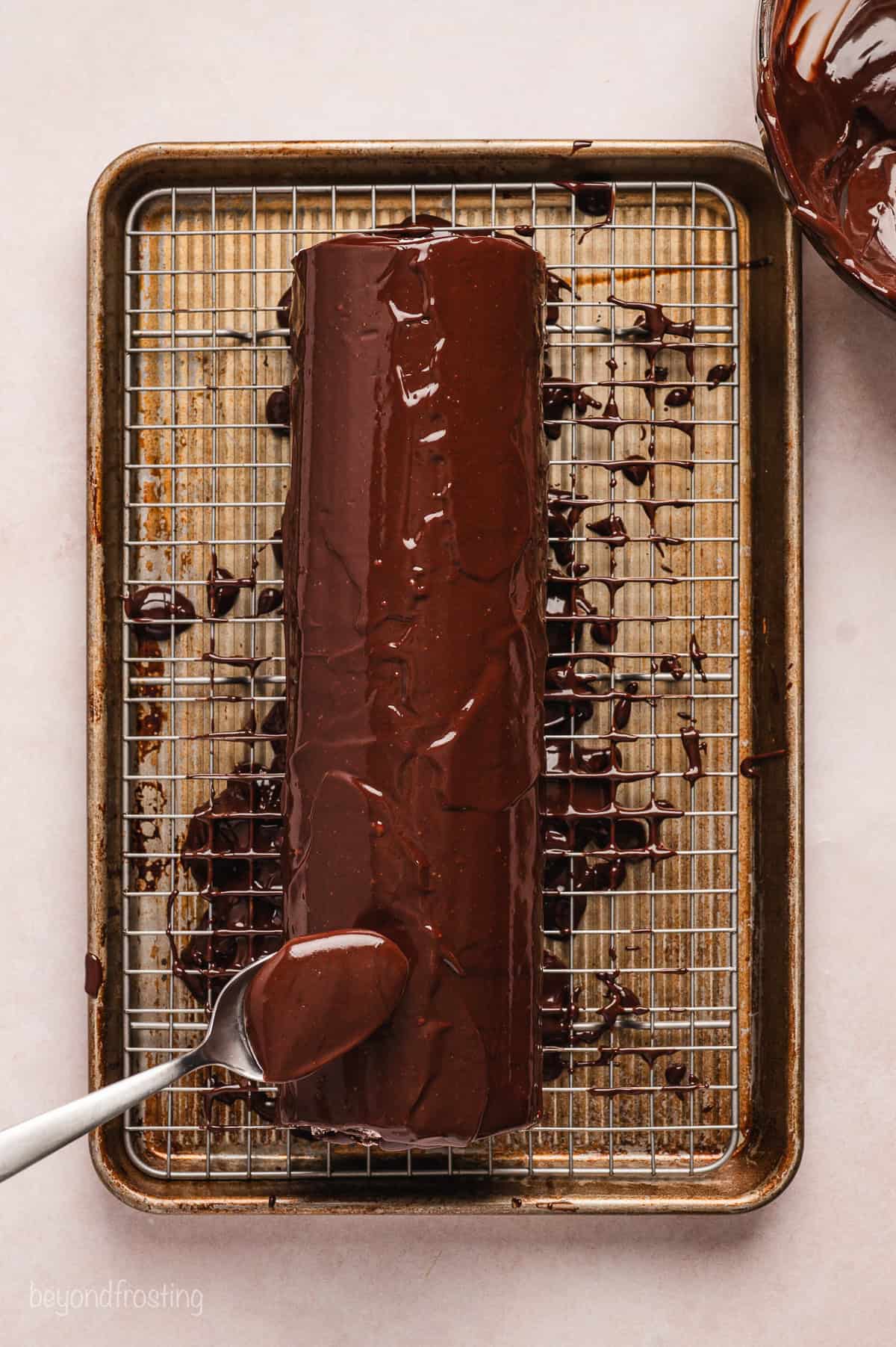 Overhead view of a spoon drizzling chocolate ganache over a chocolate cake roll on a wire rack set over a baking sheet.