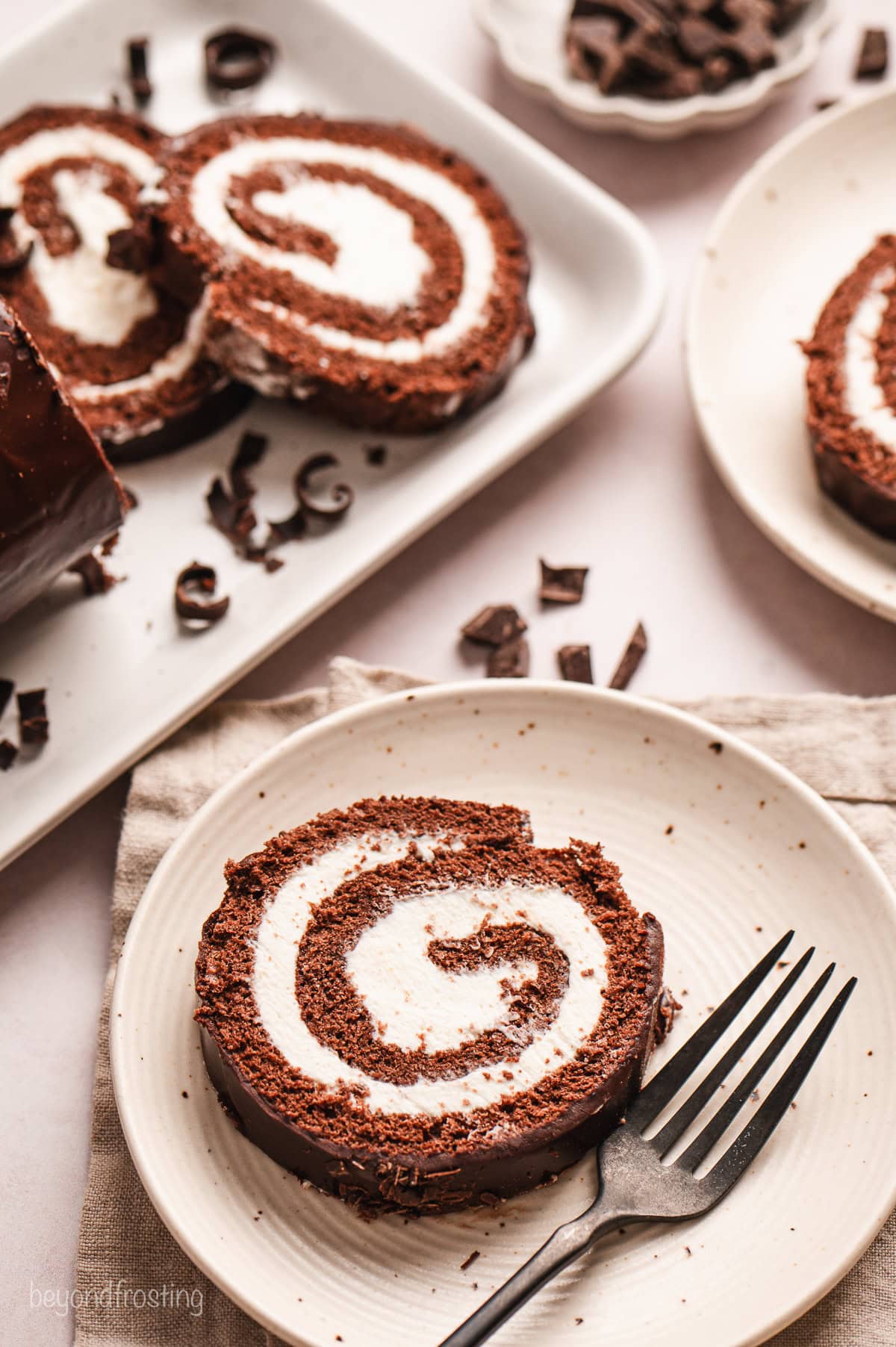 A slice of chocolate cake roll on a white plate with a fork, next to the rest of the cake with slices cut from the end.