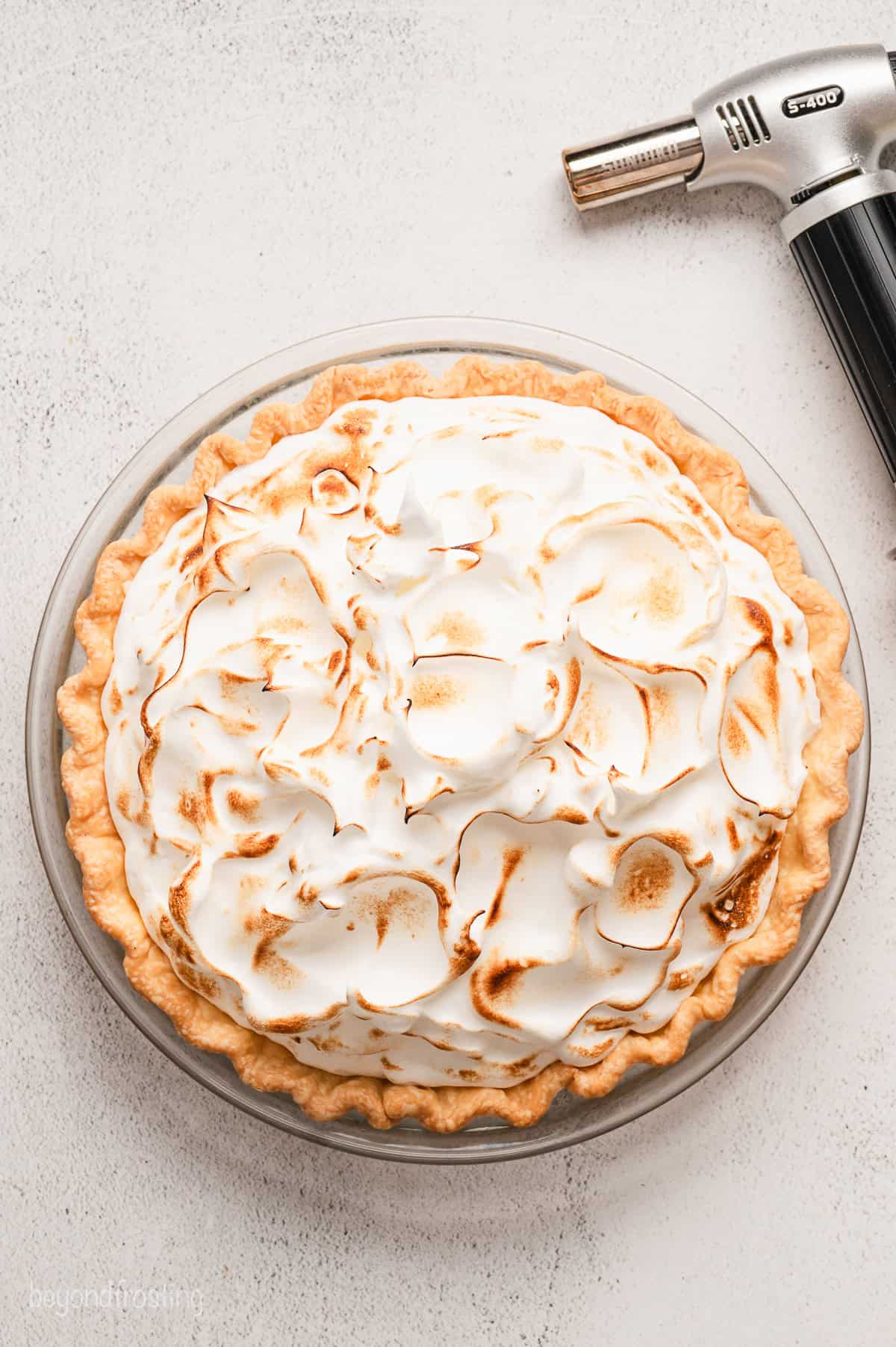 Overhead view of a lemon meringue pie topped with toasted meringue in a pie plate, next to a kitchen torch.