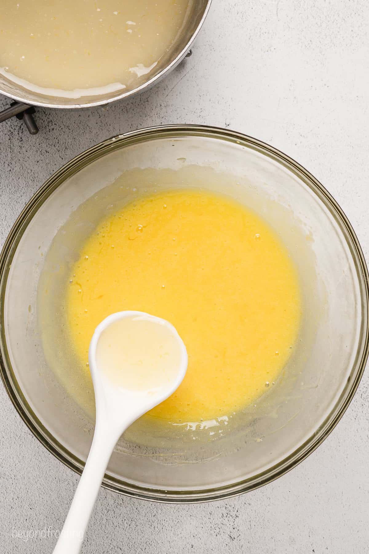 A spoon adding lemon juice mixture to a bowl of whipped egg yolks to temper them.