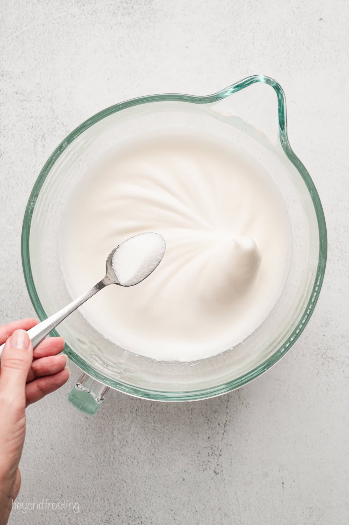 A hand adding a spoonful of salt to partially whipped meringue.